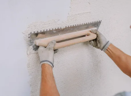 Ein Arbeiter glättet Gips an einer Wand mit einer gekerbten Kelle mit Kalkputz, wobei er sich auf eine 