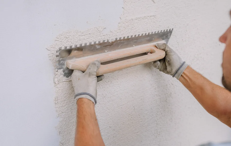 Ein Arbeiter glättet Gips an einer Wand mit einer gekerbten Kelle mit Kalkputz, wobei er sich auf eine 